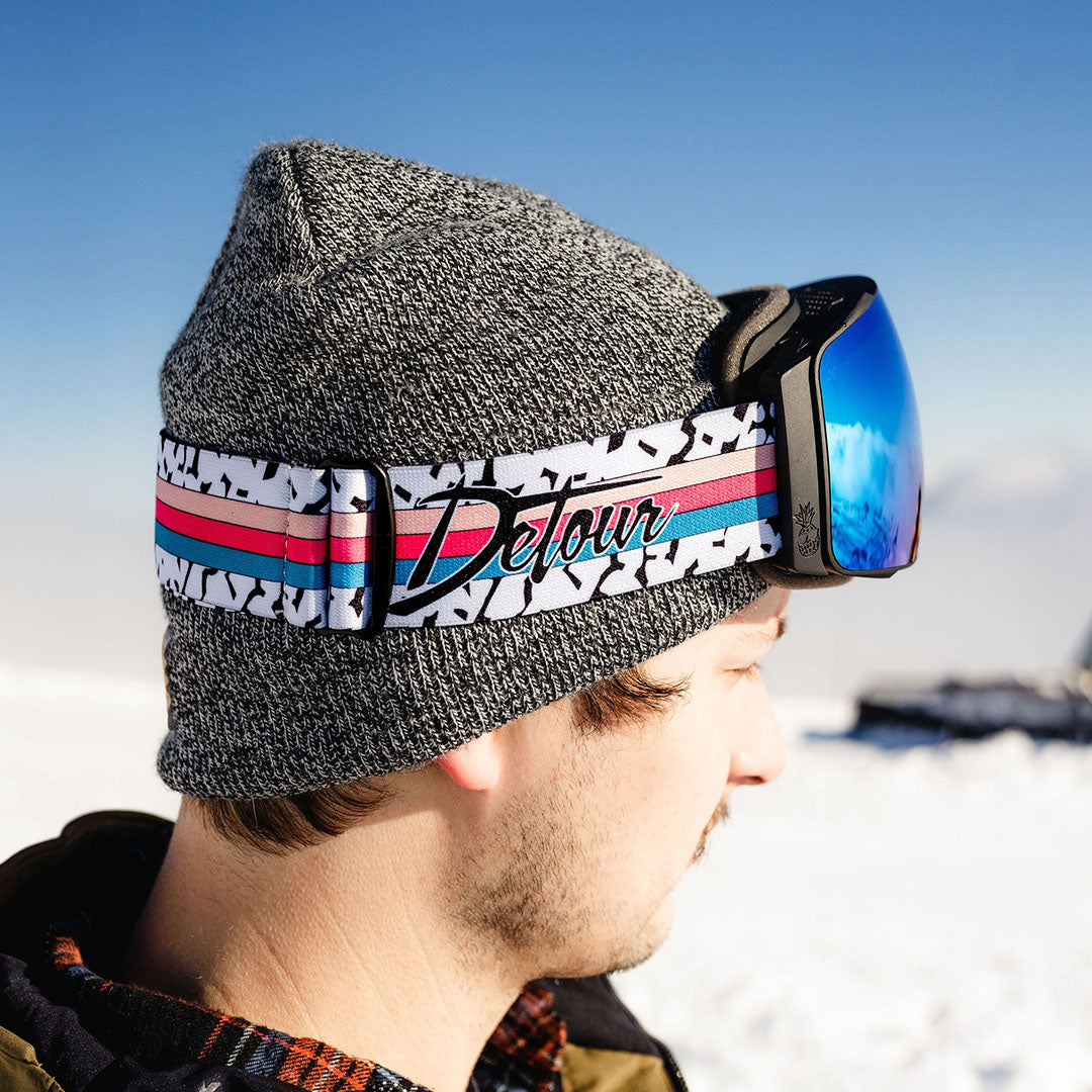 Person carrying skis in snowy environment, wearing colorful goggles and black gear.