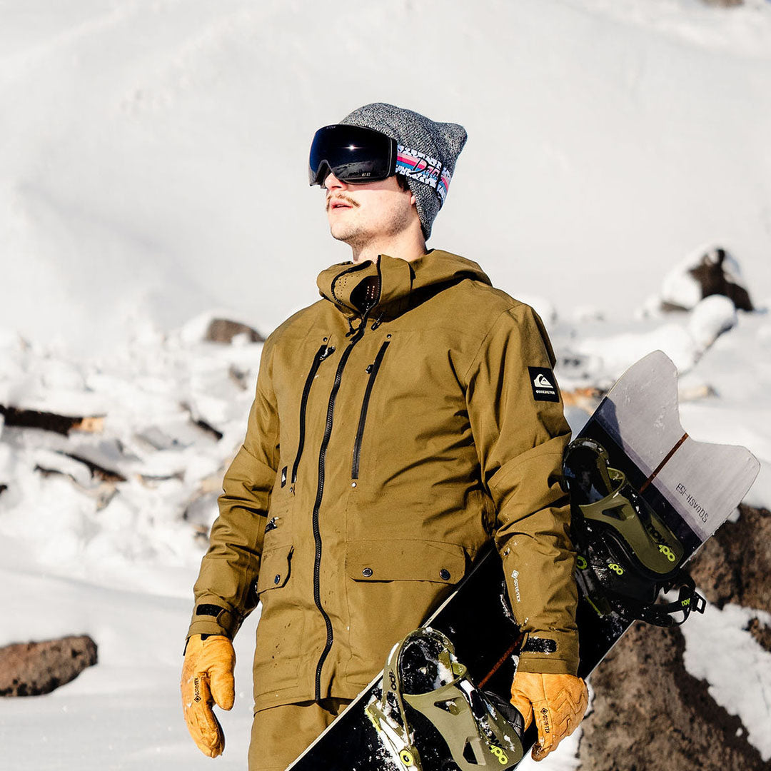 Person wearing blue jacket holding ski goggles by a lake with snowy mountains in the background.