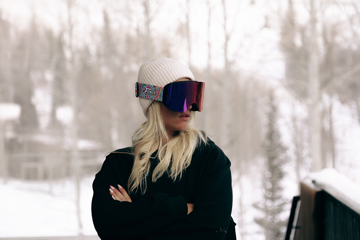 Person wearing a ski helmet and mirrored goggles, winking with snowy trees in the background.