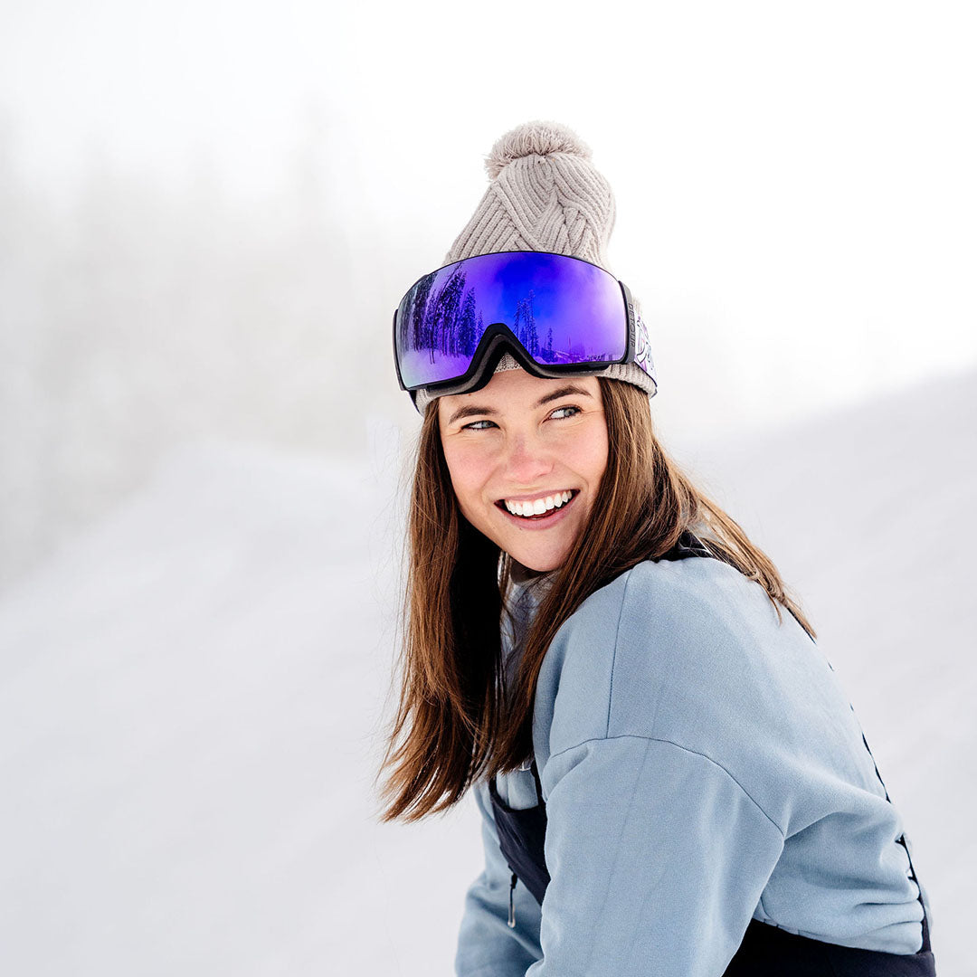 Ski goggles with a 'Detour' strap on a red snowboard, against a blue sky.